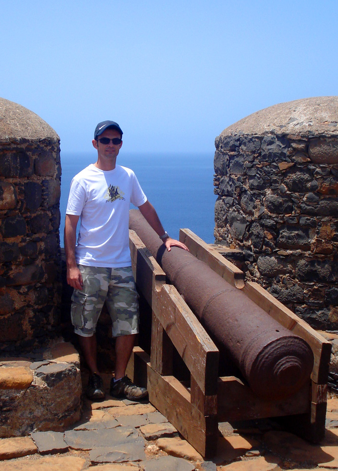 man standing next to old cannon at the edge of a hill