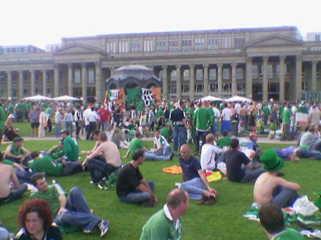 a group of people sit on grass in the sun, surrounded by a large building