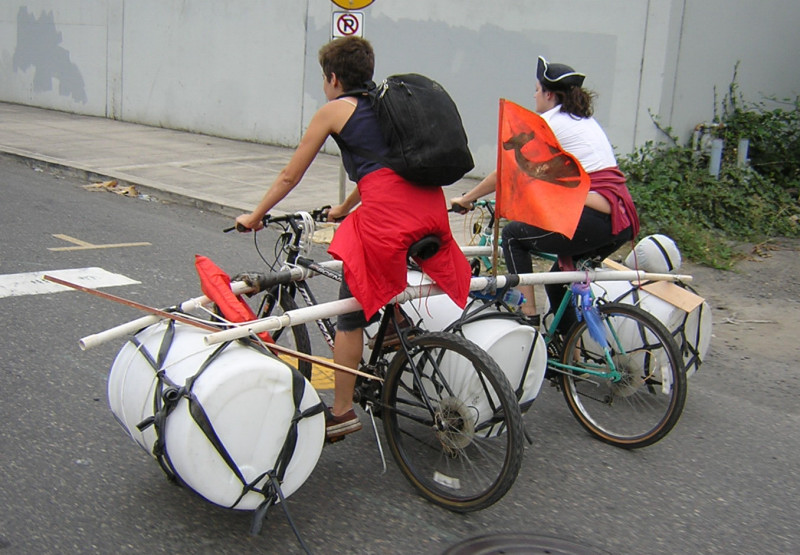 two people ride bicycles holding large drums