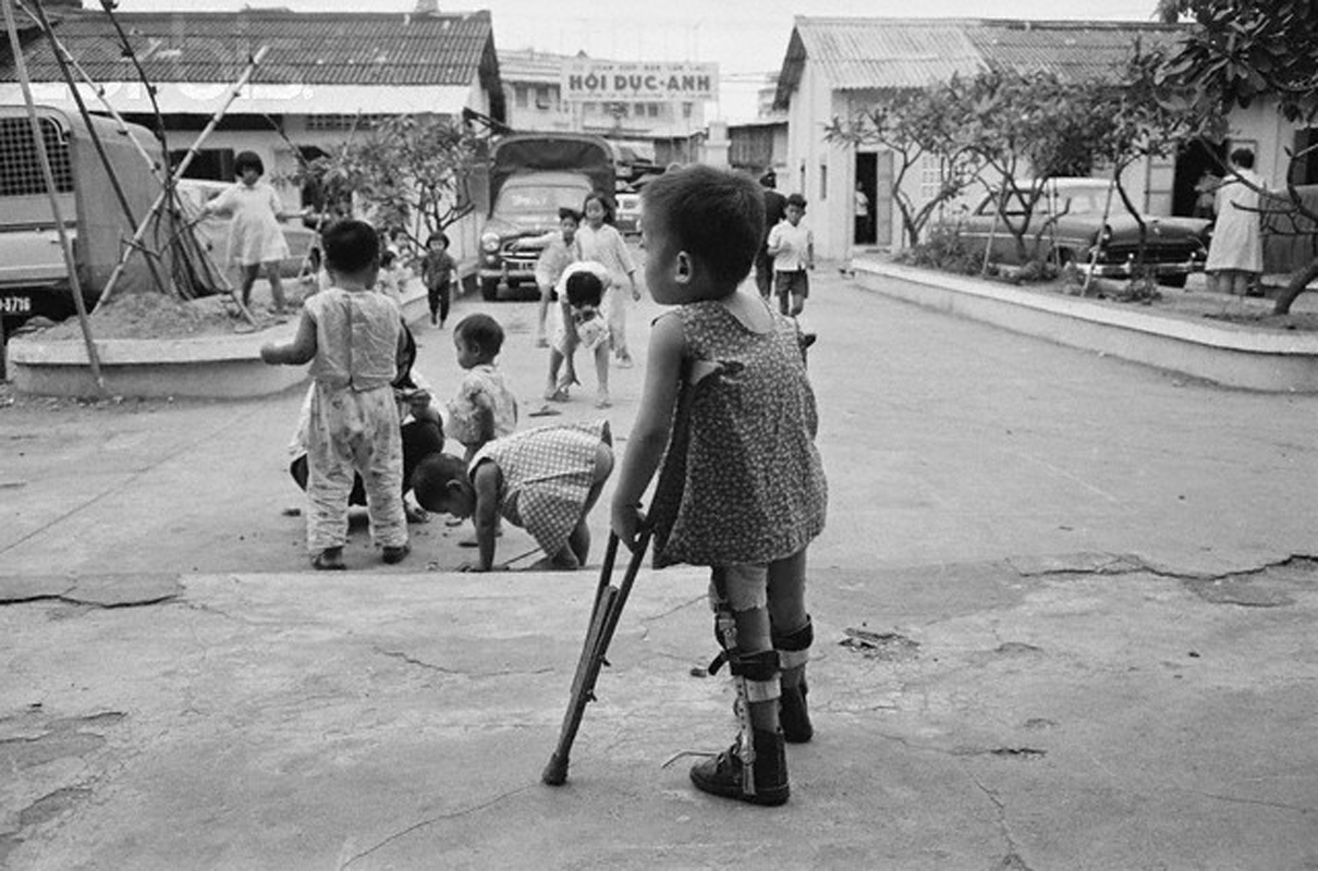 a boy holding a crutch and looking at a small group of children sitting in the dirt near the road