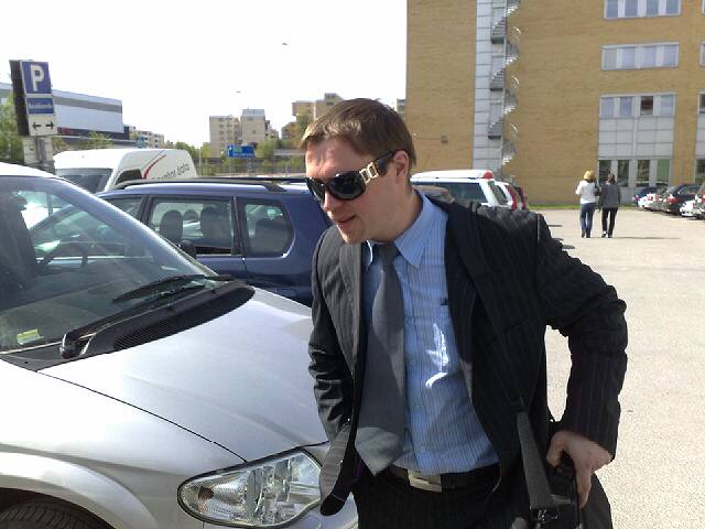 a man in suit and sunglasses with his hand on his hip, walking in a parking lot