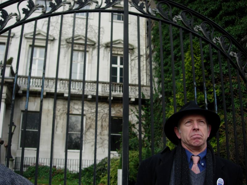 an older man wearing a suit and hat with a microphone standing next to a wrought iron gate