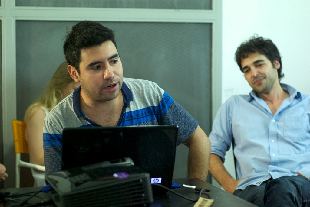 two men, one of whom is sitting on a chair and the other man is looking at a laptop