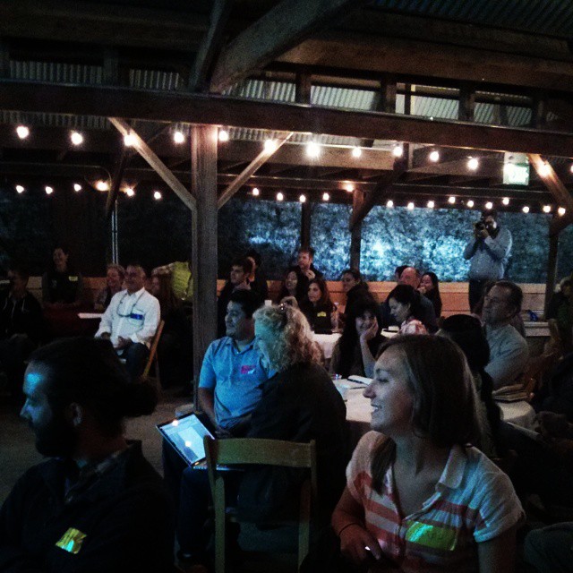 a crowd of people sit on and watch another person use their laptop