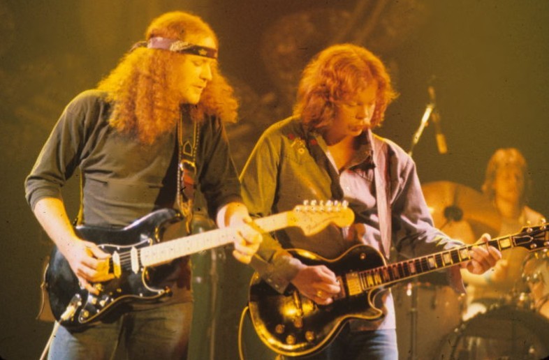 two men holding guitars on stage, one playing the guitar