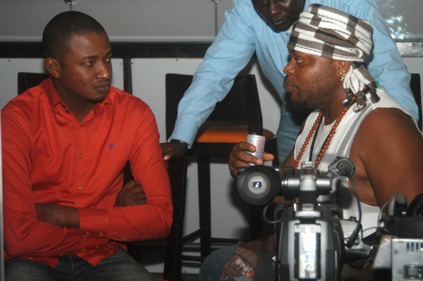 two men sit and talk together as one holds a camera