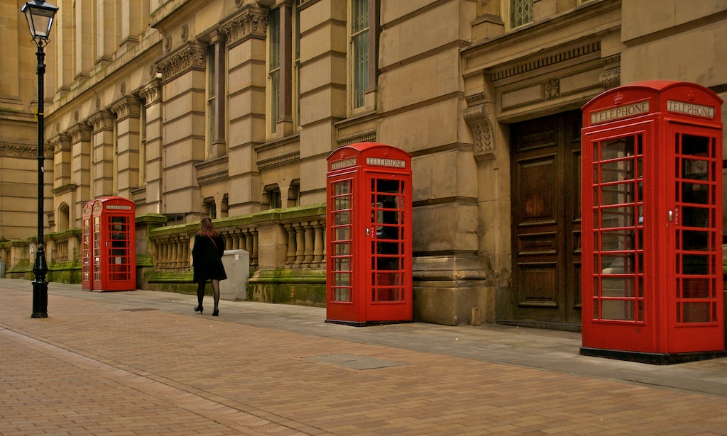 there is a man that is walking down the street with three red phone booths