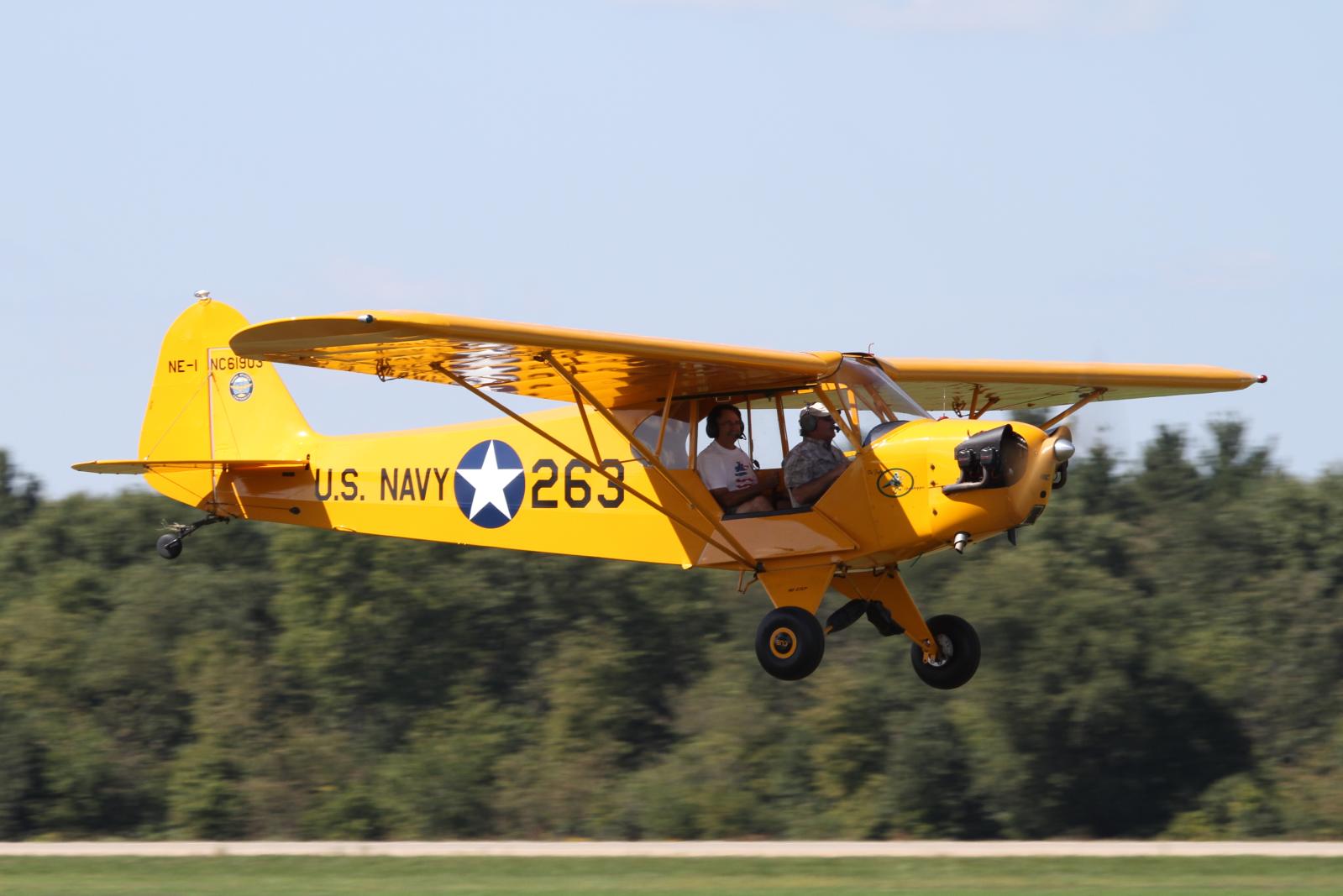 a small yellow airplane flying in front of a row of trees