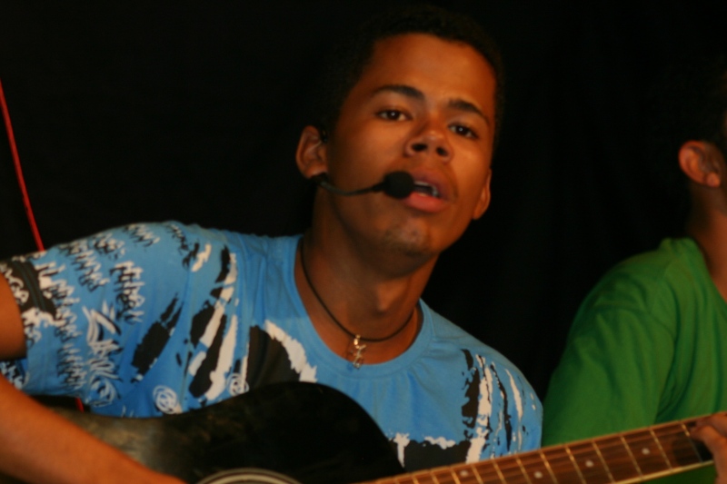 man with mustache playing guitar while another person wearing a blue shirt and green t - shirt stares off into the distance