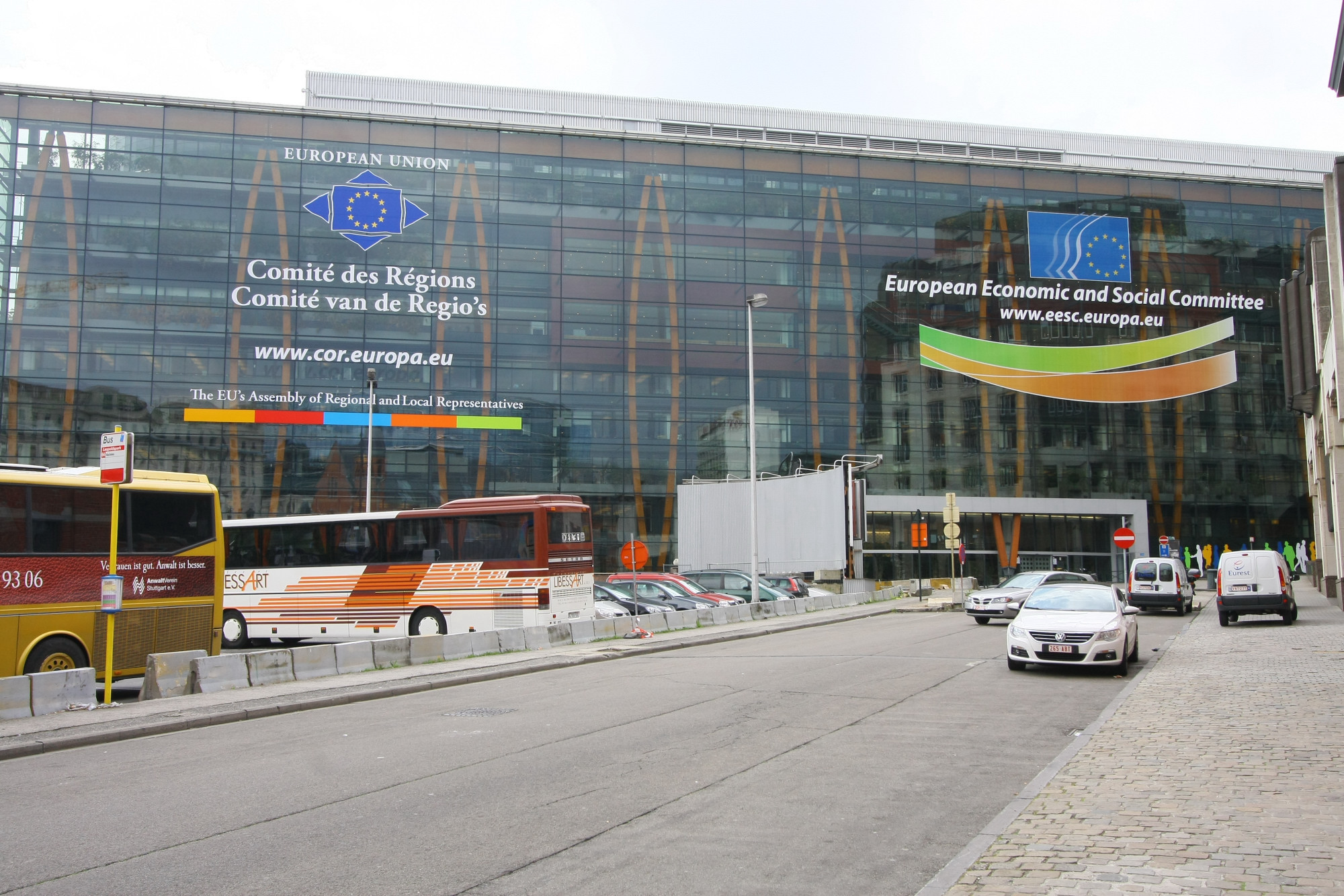 two large buses and cars are driving on the street near a large building