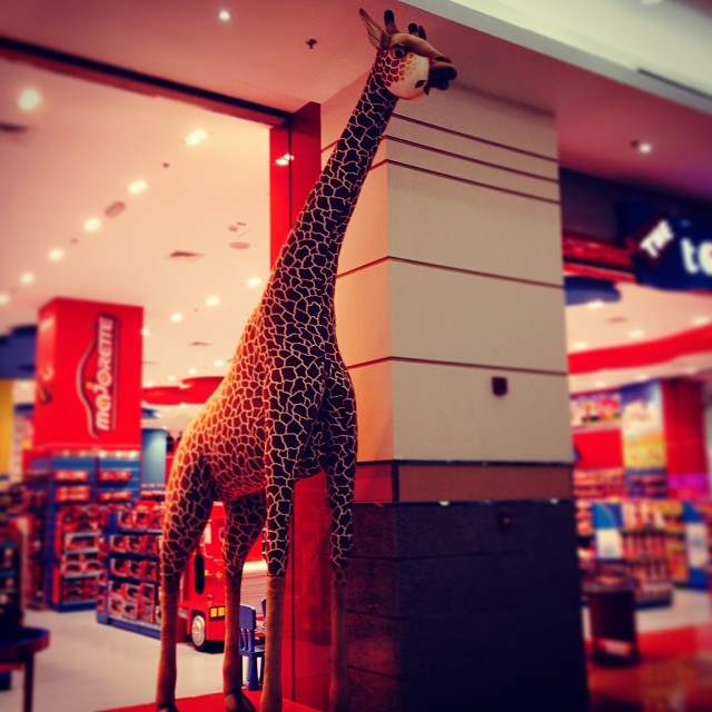giraffe statue leaning on pillar at mall with people in the background