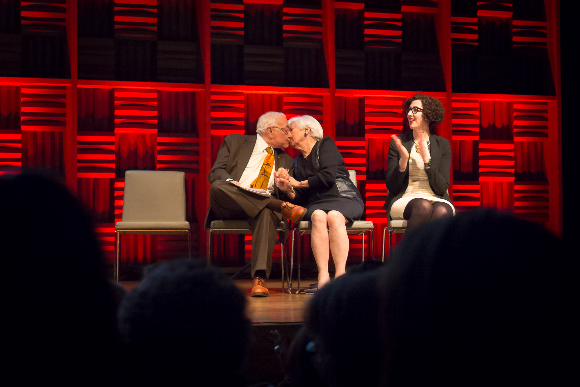 two older people seated on a stage talking to a young lady