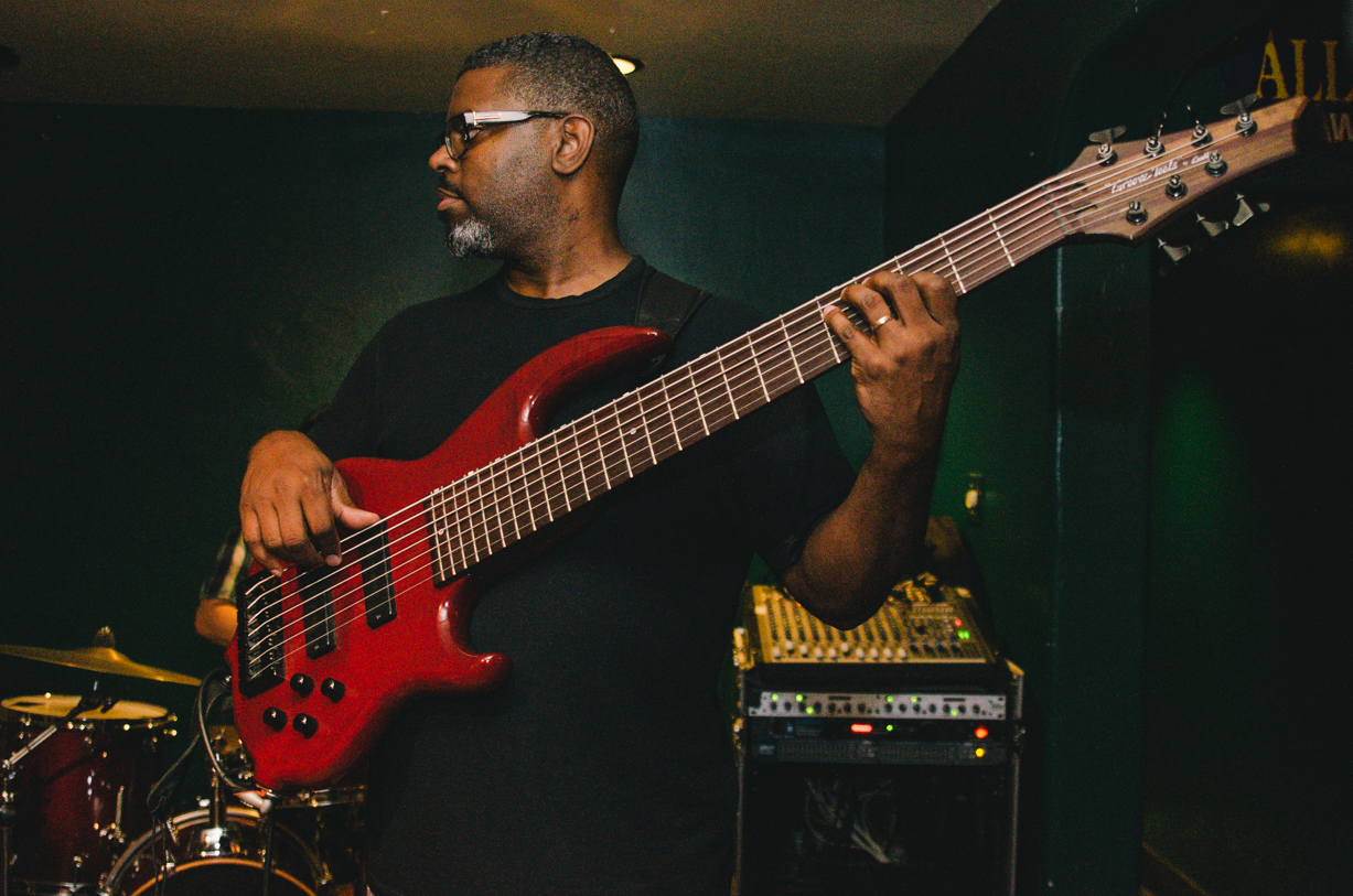 a black man with glasses holding an electric bass guitar