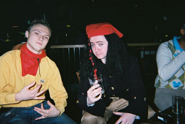 two men sitting down with drinks and one man wearing a silly hat