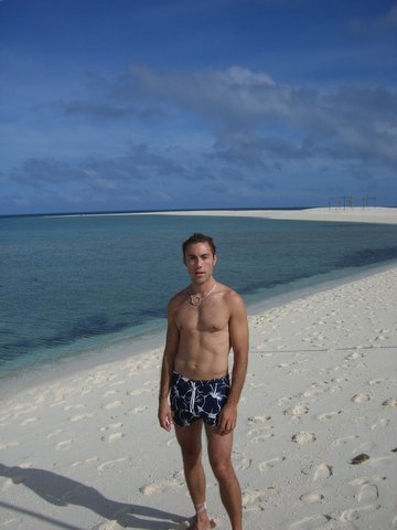 a young man in swim trunks stands on a white sandy beach by the water