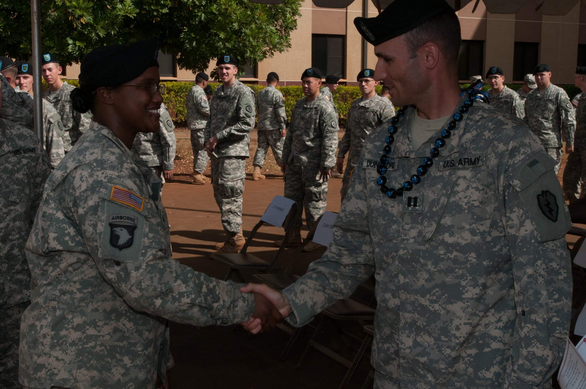 two soldiers shake hands and laugh in front of many other people