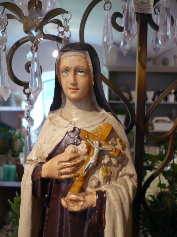 a wooden statue of an infant jesus wearing a brown and white mantle with a hand made glass chandelier behind it