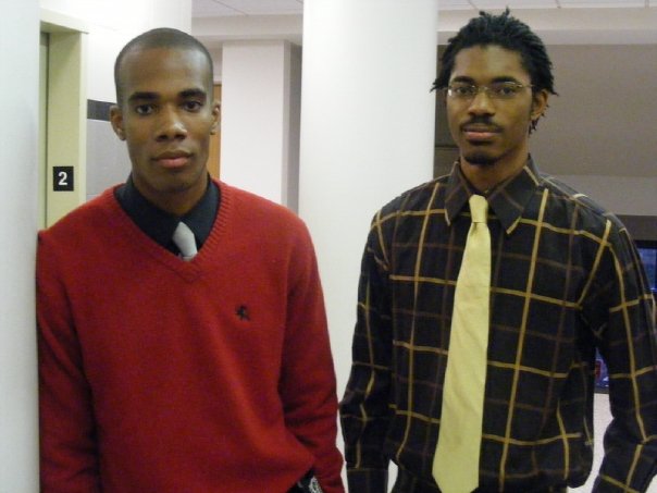 two young men stand in front of a door, one in a sweater and the other wearing a neck tie