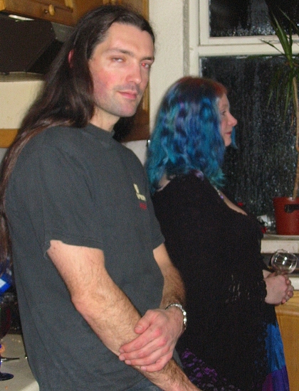 a young man with blue hair and wearing a dark shirt, standing next to his friend in the kitchen