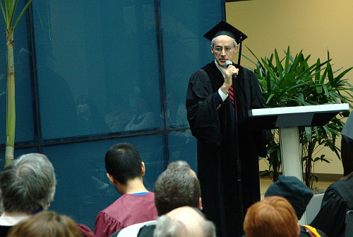 a man wearing a black graduation gown holds a microphone and speaks to people in front of him