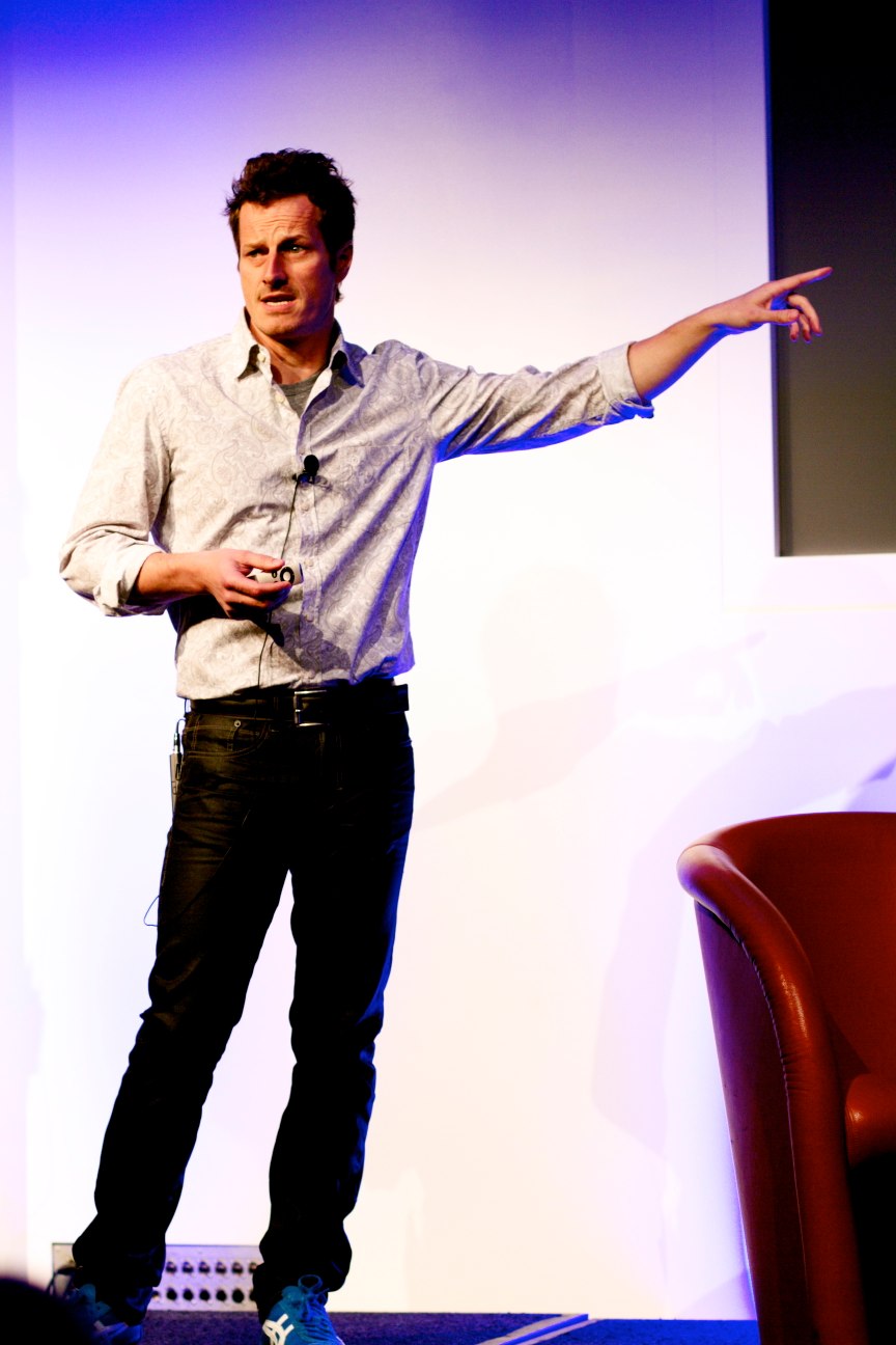 a man in a dress shirt and tie stands on stage with his arms out and gestures