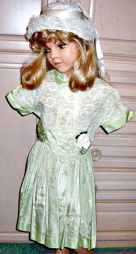 an old doll standing in front of a door wearing a green dress and a hat