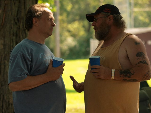 a man with tattooed arms and legs talks to another man under a tree