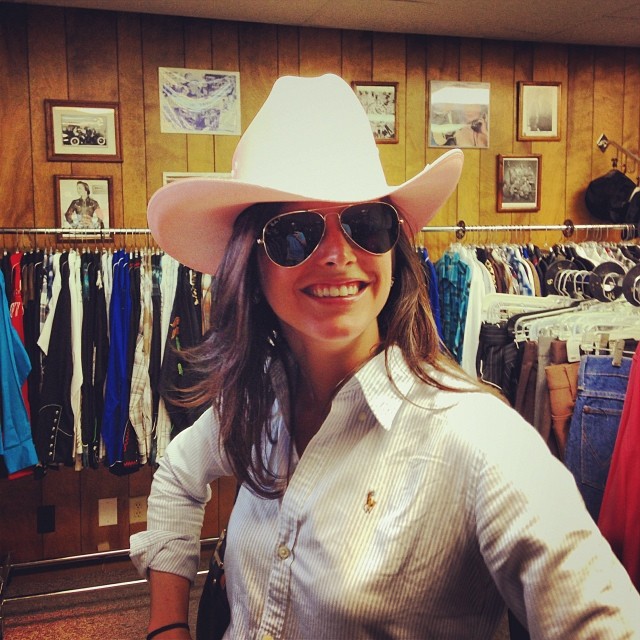 woman wearing a cowboy hat, sunglasses and smiling in a clothes shop