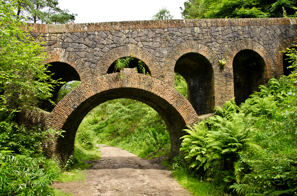 a stone structure with arches between green bushes and dirt road