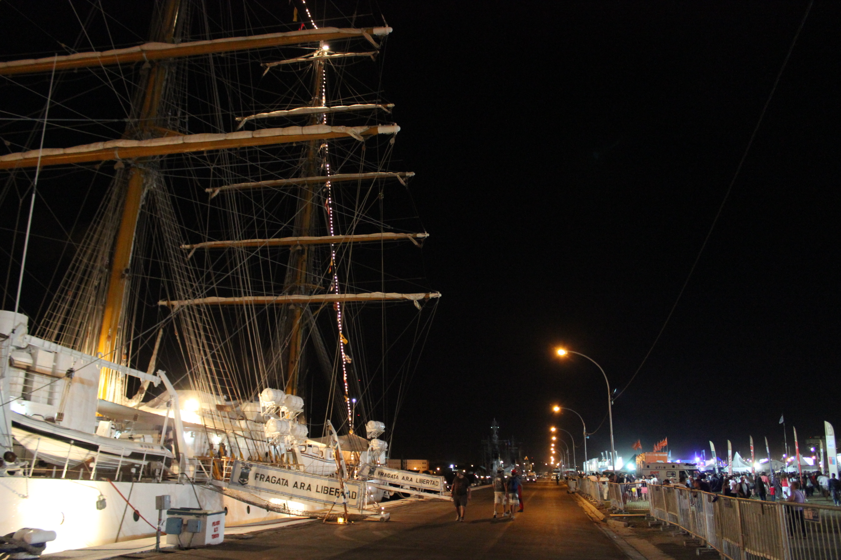 people standing on the side walk looking at a sail boat