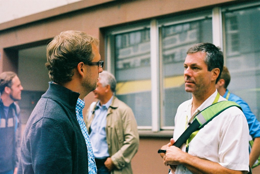 two men in conversation near another man with a camera