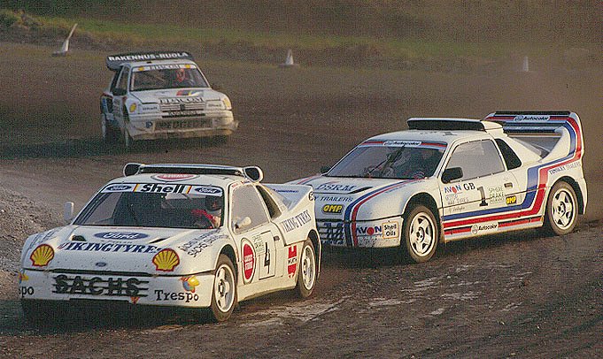two cars race across a dirt track with other cars in the background