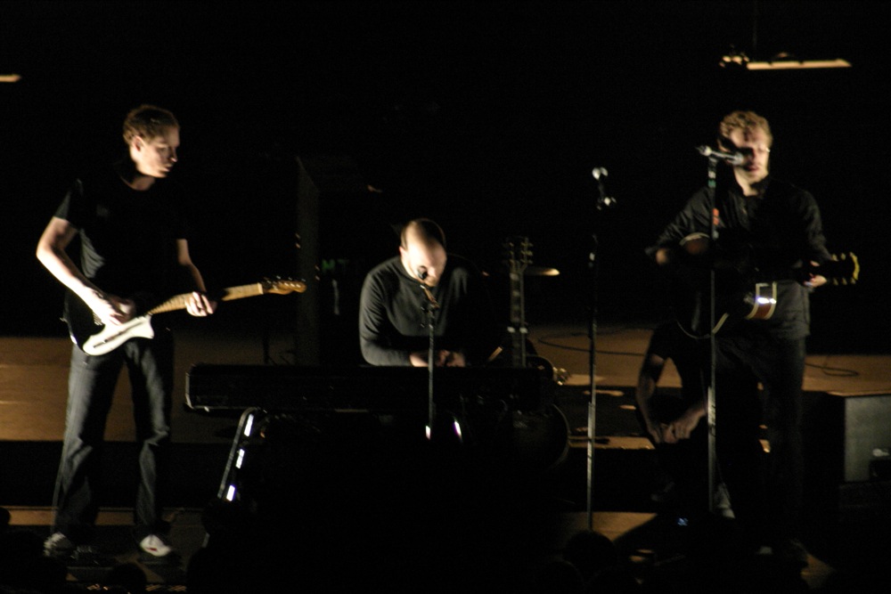 three young men stand on stage singing with guitars