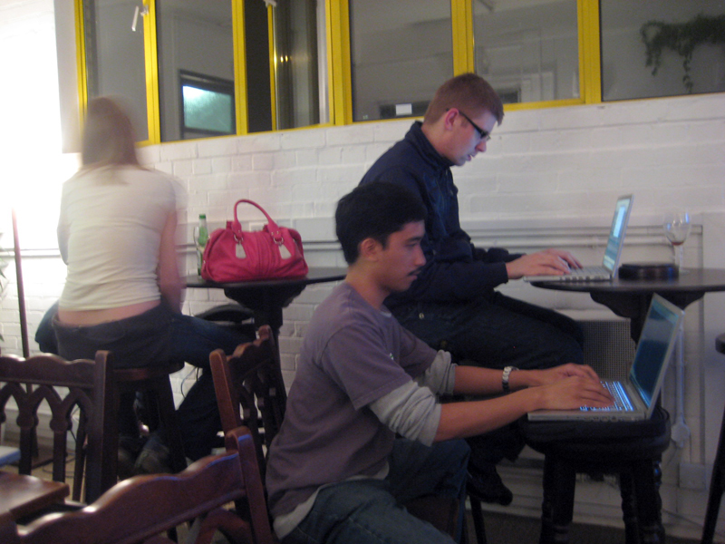 three people sit at tables with laptops on their laps