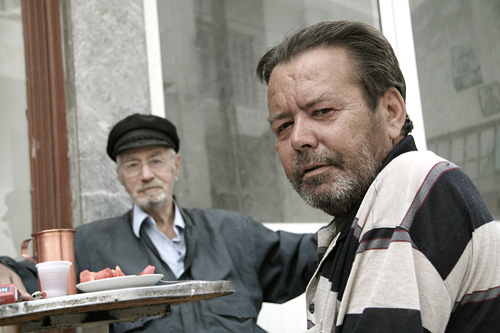a man holding a tray with food and looking at another man sitting behind it
