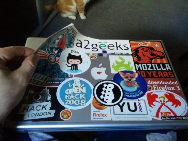 a cat standing on the floor near a laptop with stickers