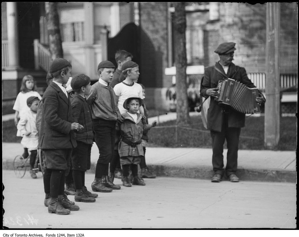 several children stand in line near a city street with an adult holding an accordo