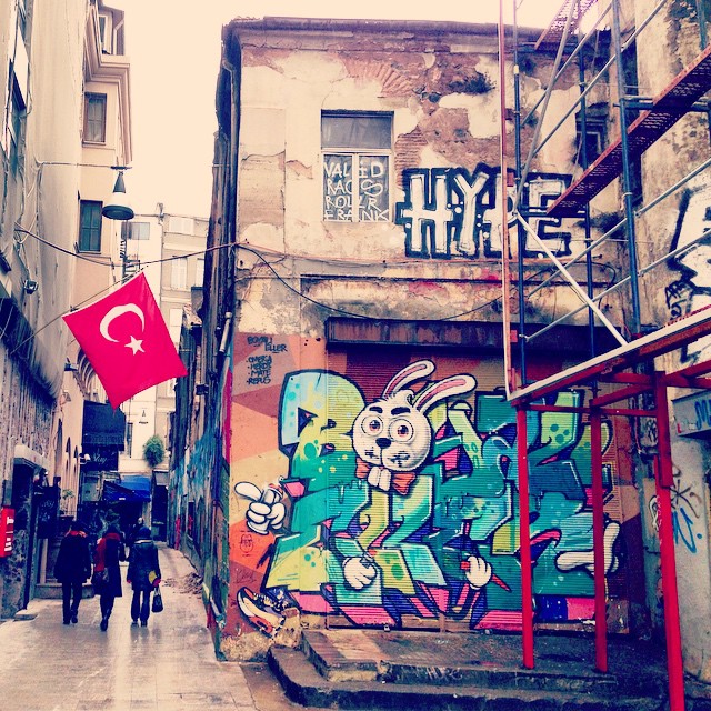 a graffiti - covered alleyway has a red flag and large paintings on the side