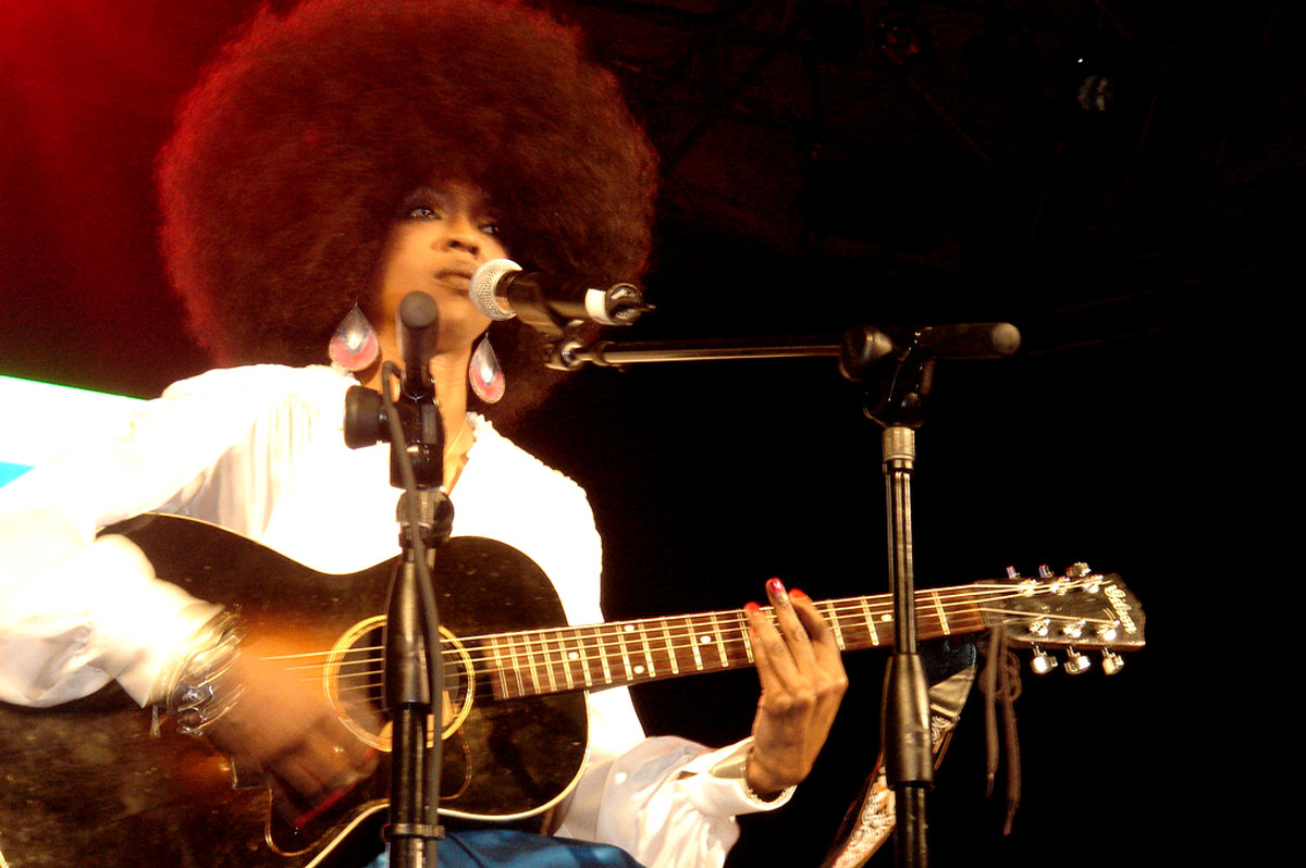 a woman with an afro sings into the microphone as she plays her guitar