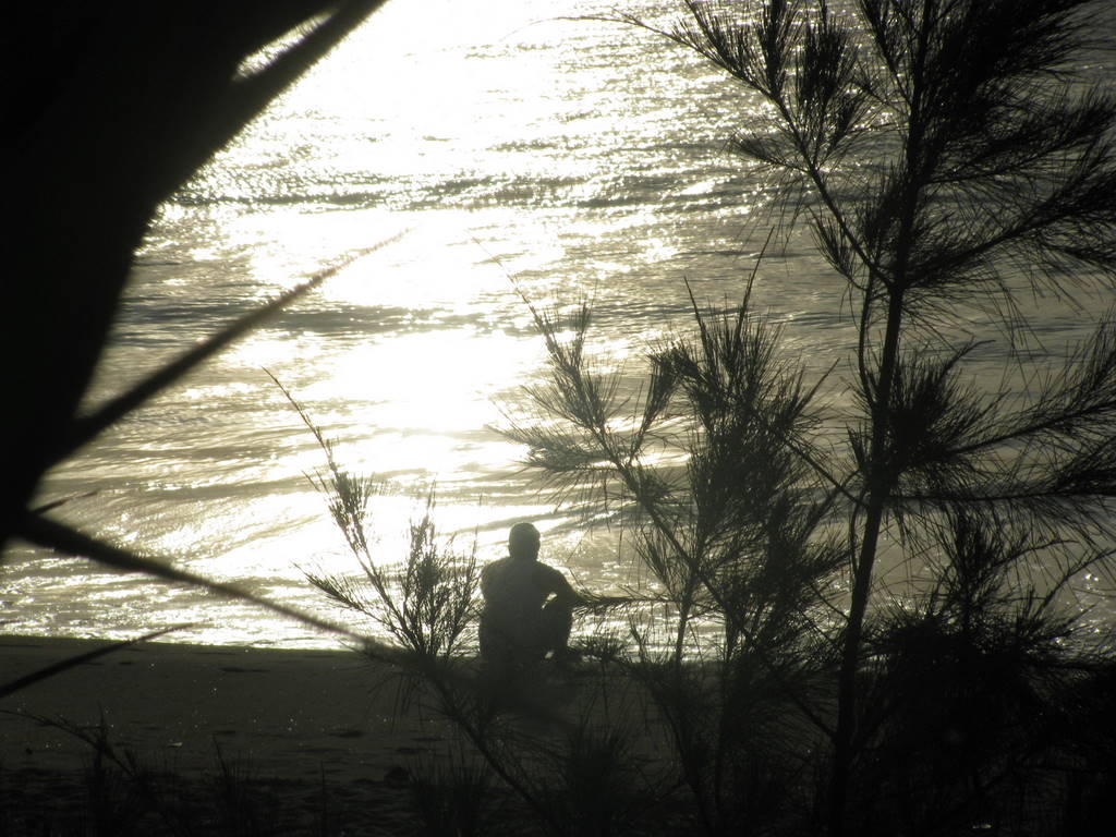 a lone man sitting on top of a beach next to water