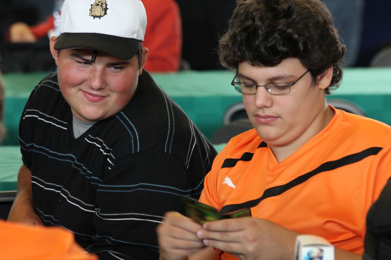 a boy in a baseball cap uses a cell phone with another person holding an item