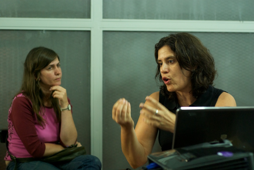 two women sit at desks in front of a laptop