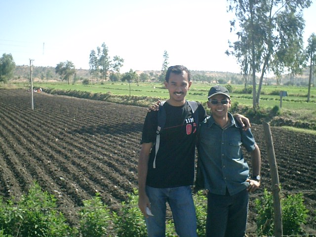 two men smile in a field while standing on a sunny day
