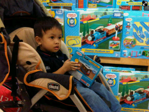 a little boy sitting in a stroller in front of a wall of toys