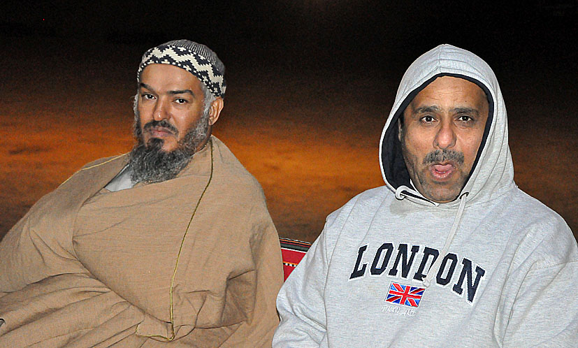 two men with a brown shawl and one wearing a gray sweatshirt and tan sweatpants, both pose for a po