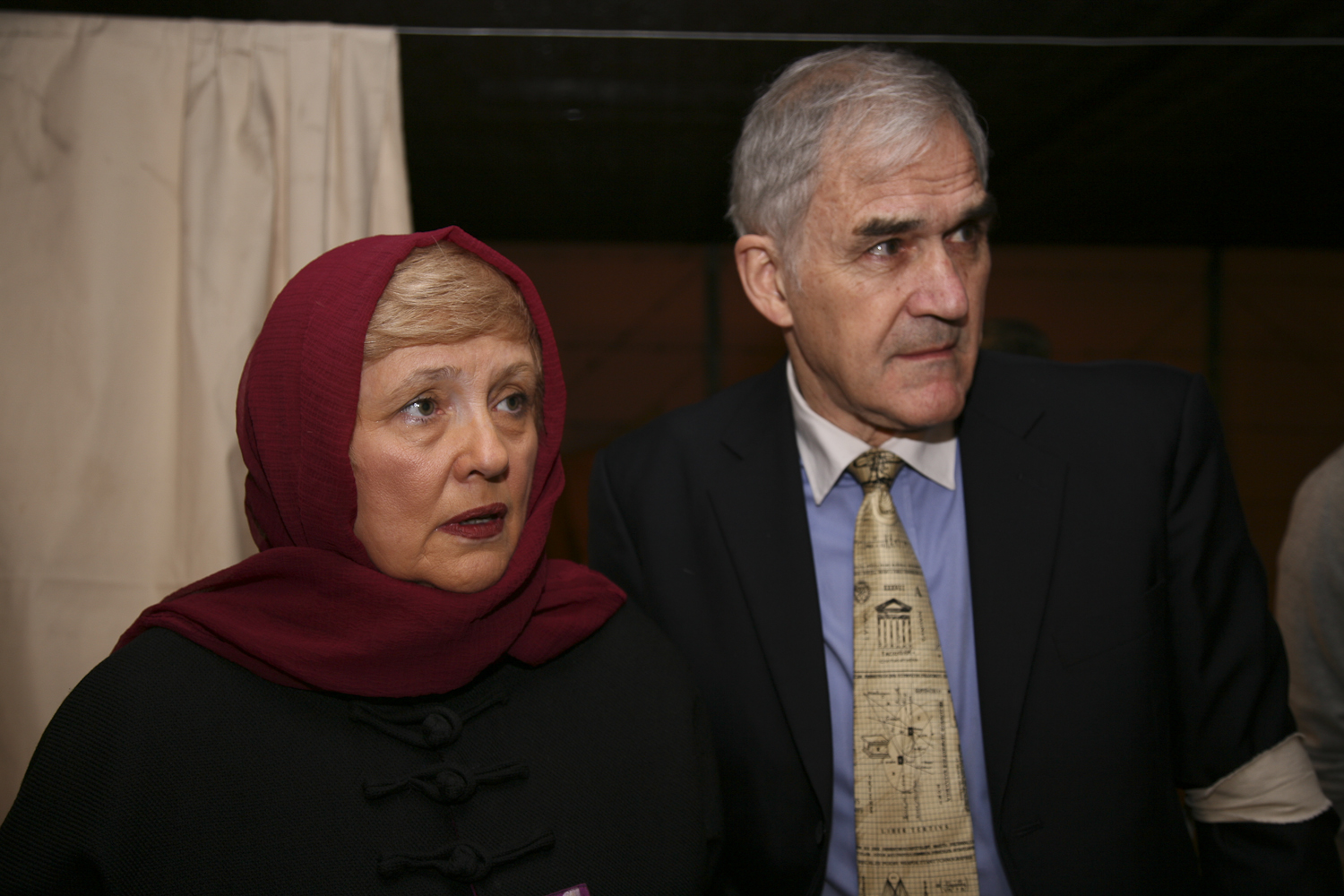 an elderly woman wearing a red head scarf standing next to a man in a suit