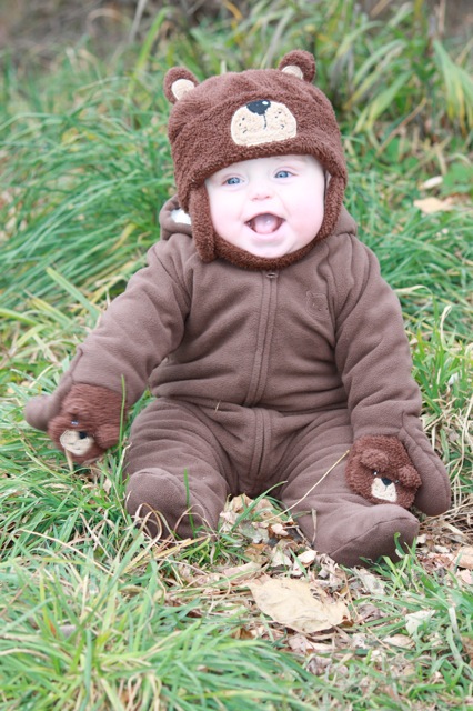 the baby wearing a brown teddy bear costume is sitting on the grass