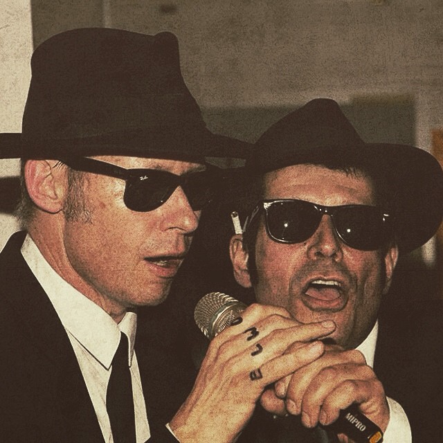 two men in black jackets and hats hold microphones