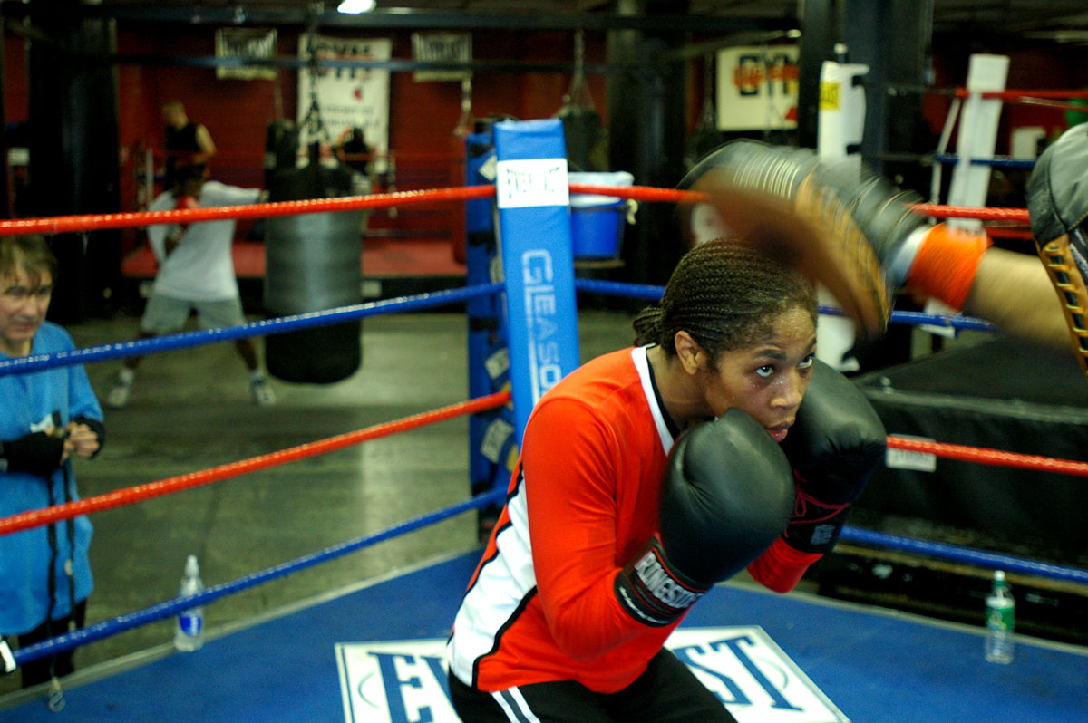 a young woman wearing boxing gloves with her trainer in the background