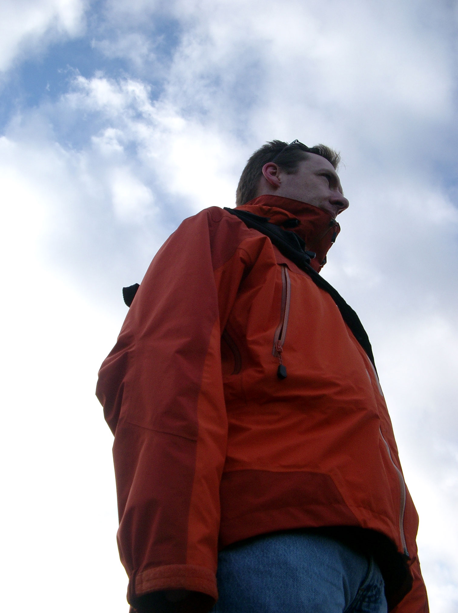 a man in an orange jacket stands against a blue cloudy sky